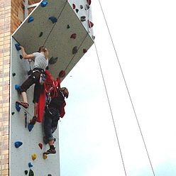 Kinder klettern mit Seilen gesichert an einer Kletterwand in großer Höhe.